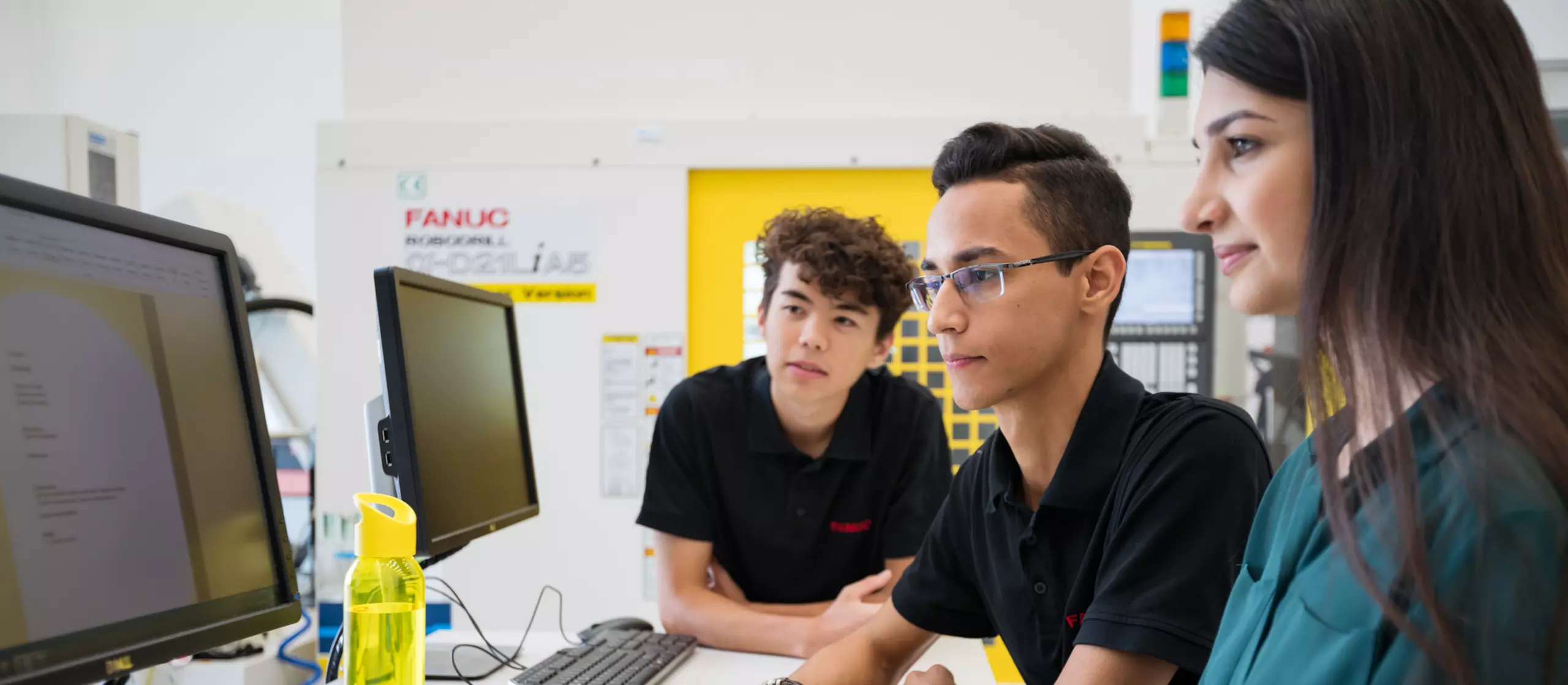 Human resources / apprentice photo with person. Three apprentices at a desk with two PCs. ROBODRILL in the background. Landscape. High resolution.