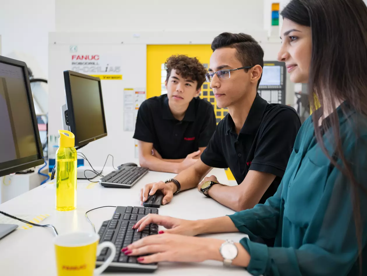 Human resources / apprentice photo with person. Three apprentices at a desk with two PCs. ROBODRILL in the background. Landscape. High resolution.